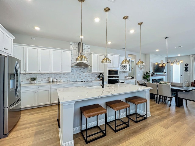 kitchen with sink, wall chimney range hood, stainless steel appliances, an island with sink, and decorative light fixtures