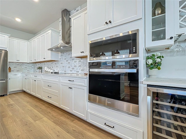 kitchen with appliances with stainless steel finishes, white cabinets, beverage cooler, wall chimney exhaust hood, and light wood-type flooring