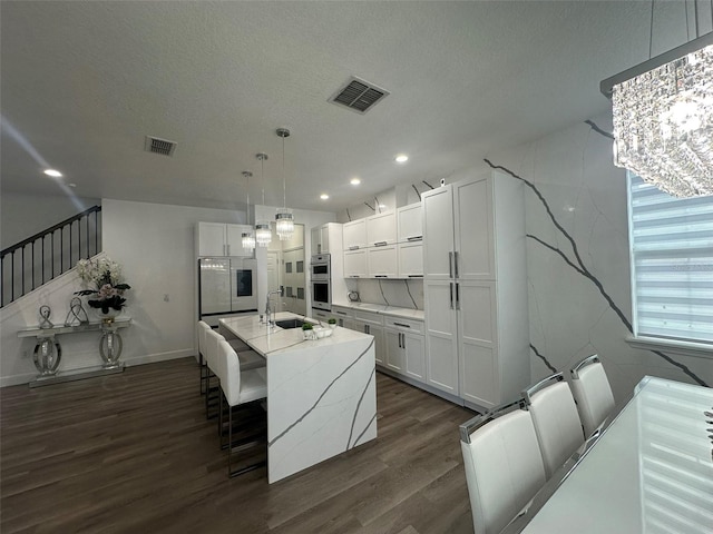 kitchen with dark wood-type flooring, hanging light fixtures, a center island with sink, light stone countertops, and white cabinets