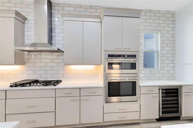 kitchen featuring wall chimney range hood, white cabinets, stainless steel appliances, beverage cooler, and backsplash