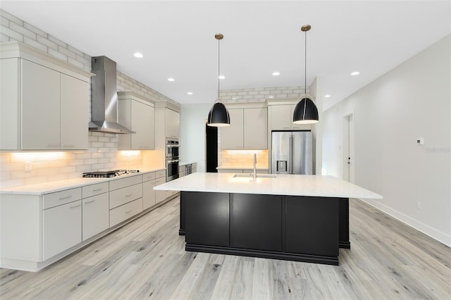 kitchen featuring sink, hanging light fixtures, a large island with sink, stainless steel appliances, and wall chimney range hood