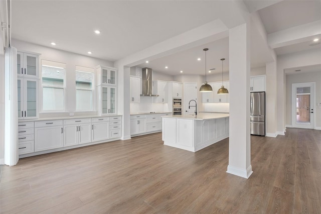 kitchen featuring wall chimney exhaust hood, white cabinetry, hanging light fixtures, stainless steel appliances, and a kitchen island with sink