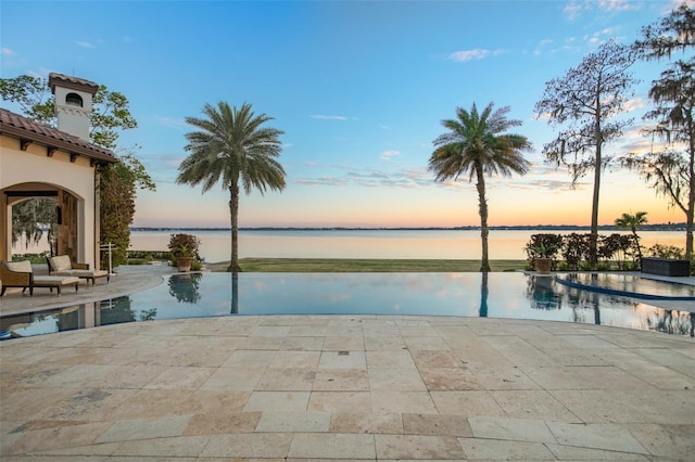 pool at dusk featuring an infinity pool, a patio, and a water view