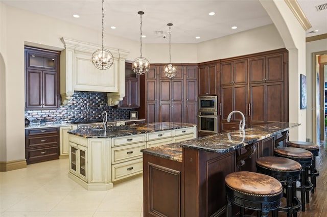 kitchen with an island with sink, backsplash, cream cabinets, arched walkways, and appliances with stainless steel finishes