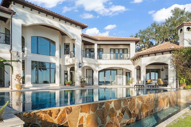 back of property with stucco siding, a balcony, an infinity pool, and a patio area