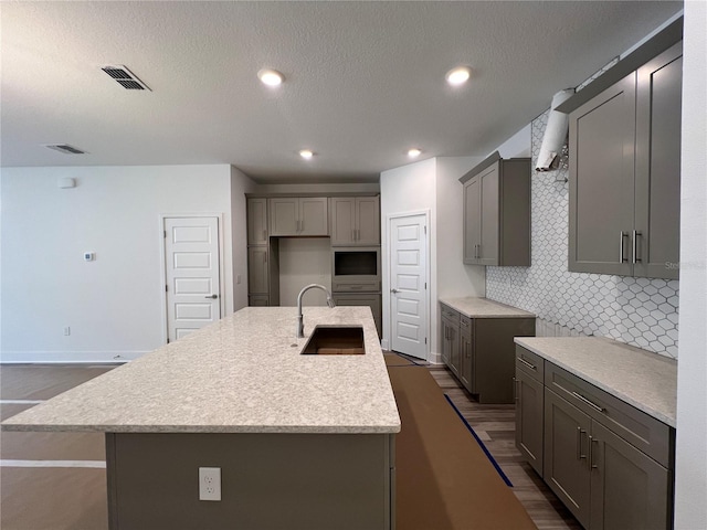 kitchen with visible vents, a kitchen island with sink, gray cabinetry, a sink, and backsplash