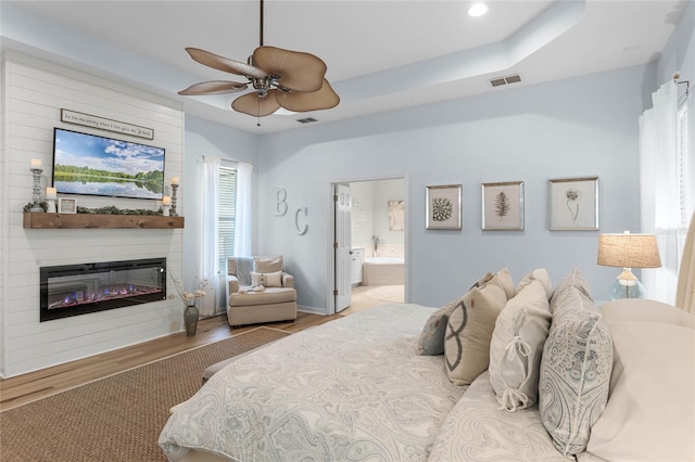 bedroom featuring a large fireplace, ceiling fan, a tray ceiling, light hardwood / wood-style floors, and ensuite bath