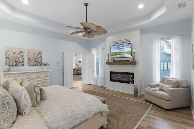 bedroom with a fireplace, a tray ceiling, light hardwood / wood-style flooring, and ceiling fan