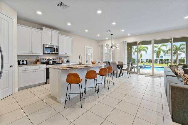 kitchen with appliances with stainless steel finishes, a kitchen island with sink, white cabinetry, light stone counters, and decorative light fixtures