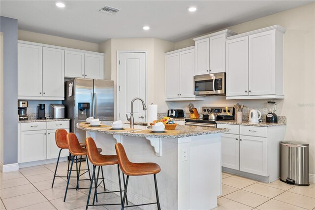 kitchen with light tile patterned flooring, white cabinetry, light stone counters, stainless steel appliances, and a center island with sink