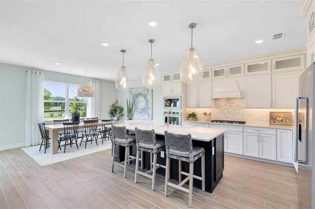 kitchen with decorative light fixtures, white cabinetry, decorative backsplash, stainless steel appliances, and a center island with sink