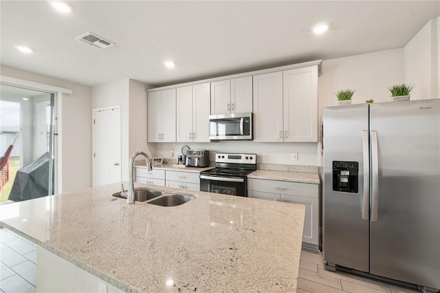 kitchen with stainless steel appliances, visible vents, a sink, an island with sink, and light stone countertops