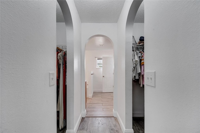 corridor featuring baseboards, wood finished floors, a textured wall, arched walkways, and a textured ceiling