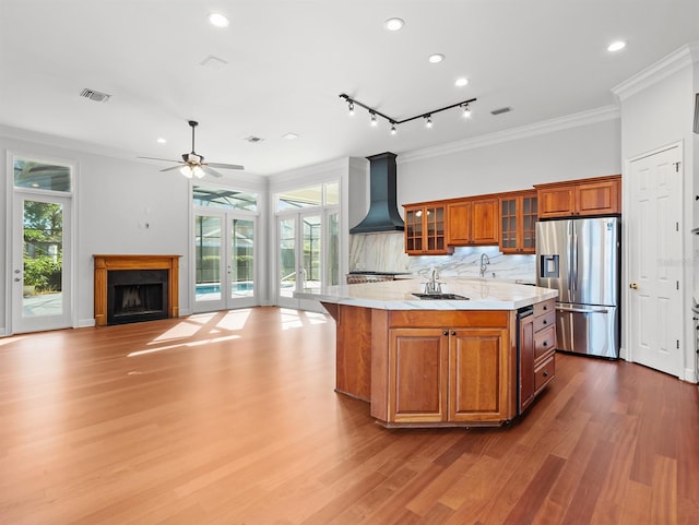 kitchen featuring premium range hood, sink, crown molding, stainless steel fridge with ice dispenser, and a kitchen island with sink