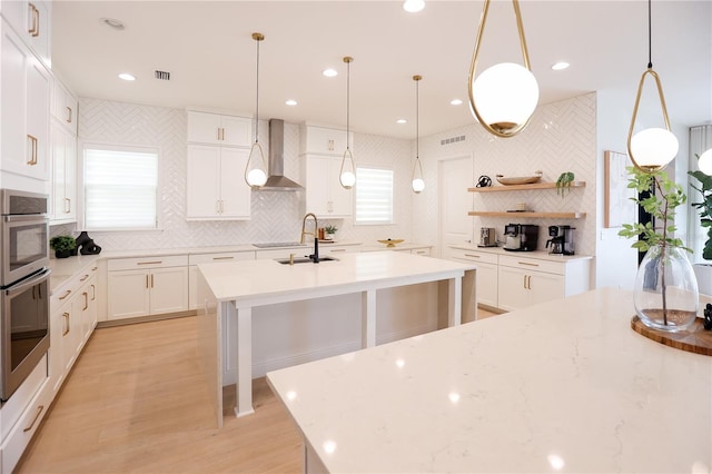 kitchen with decorative light fixtures, white cabinetry, backsplash, a kitchen island with sink, and wall chimney range hood