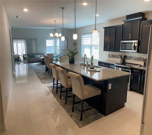 kitchen with sink, light tile patterned floors, stainless steel appliances, light stone countertops, and an island with sink