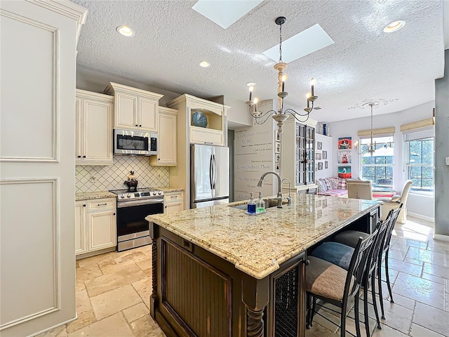 kitchen with pendant lighting, appliances with stainless steel finishes, an inviting chandelier, light stone countertops, and an island with sink