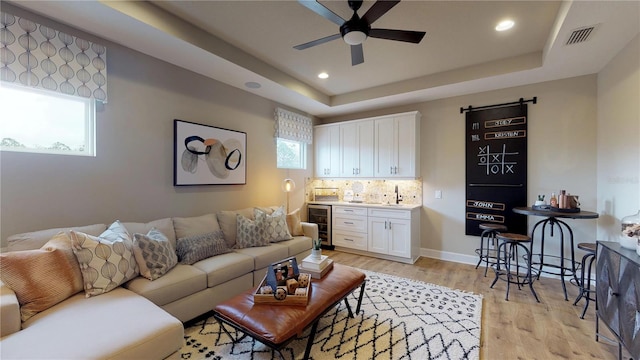 living room featuring wine cooler, wet bar, a raised ceiling, ceiling fan, and light hardwood / wood-style floors