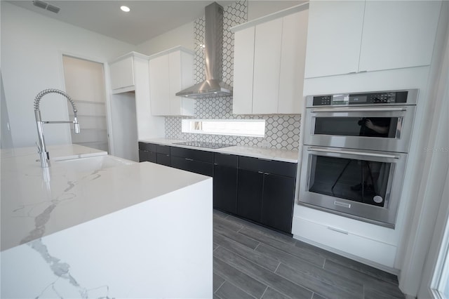 kitchen with stainless steel double oven, a sink, white cabinets, wall chimney range hood, and tasteful backsplash