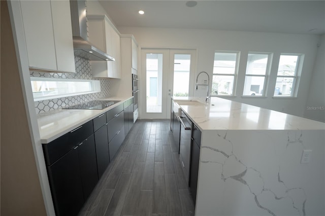 kitchen with tasteful backsplash, black electric stovetop, a large island with sink, wall chimney exhaust hood, and a sink