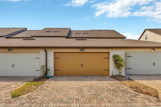 view of front of house with community garages, stucco siding, a shingled roof, and decorative driveway