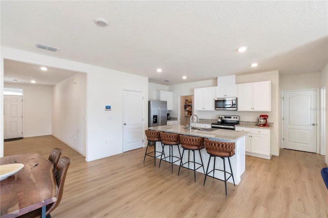 kitchen featuring a kitchen island with sink, white cabinetry, stainless steel appliances, light stone counters, and a kitchen bar