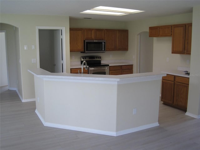 kitchen with arched walkways, brown cabinets, a center island with sink, light wood finished floors, and stainless steel appliances