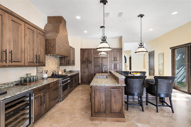 kitchen with sink, custom exhaust hood, high end range, a center island with sink, and beverage cooler