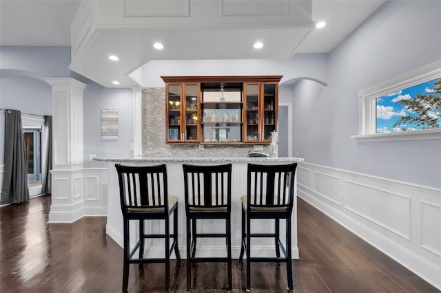 bar with ornate columns, light stone countertops, dark wood-type flooring, and backsplash