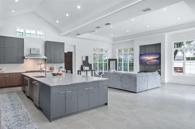 kitchen featuring gray cabinets, high vaulted ceiling, plenty of natural light, a center island with sink, and wall chimney exhaust hood