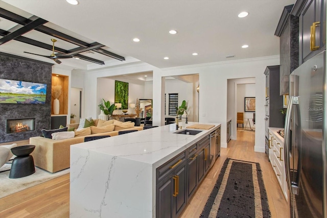 kitchen featuring sink, a center island with sink, stainless steel refrigerator, a large fireplace, and light stone countertops