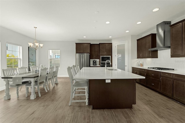 kitchen with an island with sink, sink, hanging light fixtures, stainless steel appliances, and wall chimney range hood