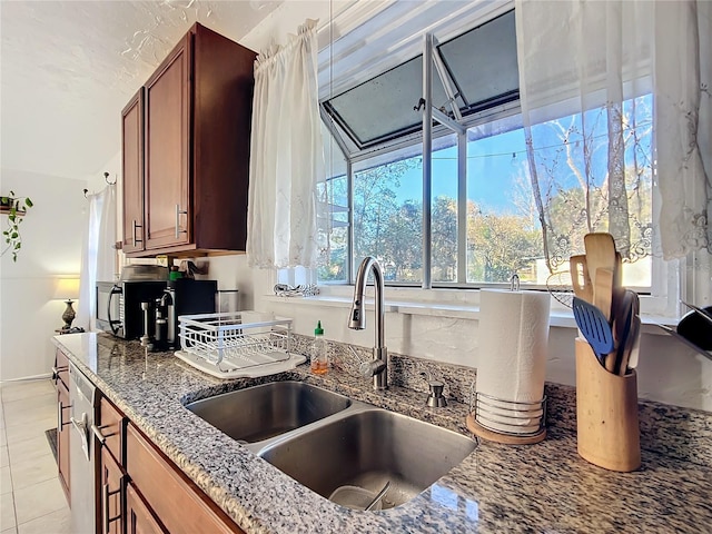 kitchen with light stone counters, light tile patterned floors, stainless steel dishwasher, brown cabinetry, and a sink