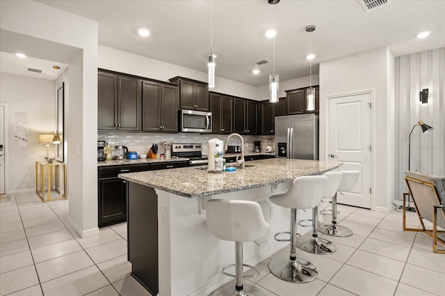 kitchen with sink, light stone counters, an island with sink, pendant lighting, and stainless steel appliances