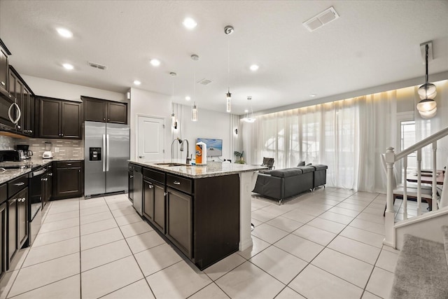 kitchen with an island with sink, sink, hanging light fixtures, stainless steel appliances, and light stone countertops