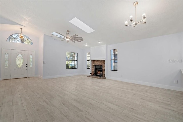 unfurnished living room featuring lofted ceiling with skylight, ceiling fan with notable chandelier, a brick fireplace, and light wood-type flooring