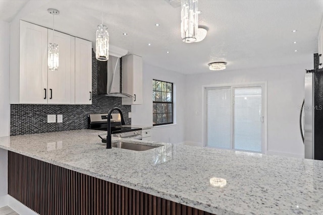 kitchen with wall chimney range hood, sink, light stone counters, white cabinets, and decorative light fixtures