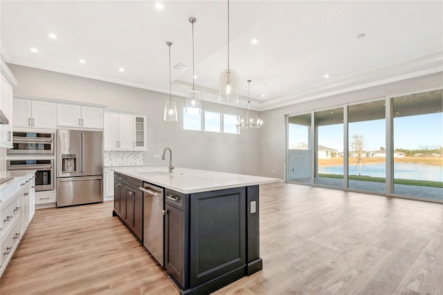 kitchen with a water view, appliances with stainless steel finishes, pendant lighting, a kitchen island with sink, and white cabinets