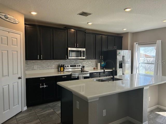 kitchen with sink, a textured ceiling, appliances with stainless steel finishes, a kitchen island with sink, and backsplash