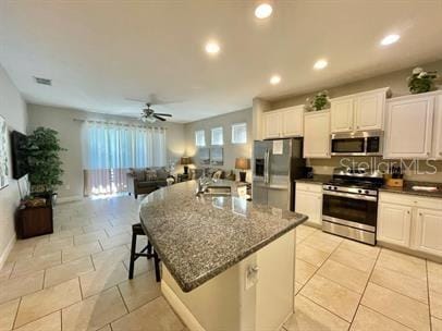 kitchen with light tile patterned flooring, appliances with stainless steel finishes, white cabinetry, dark stone counters, and a kitchen island with sink