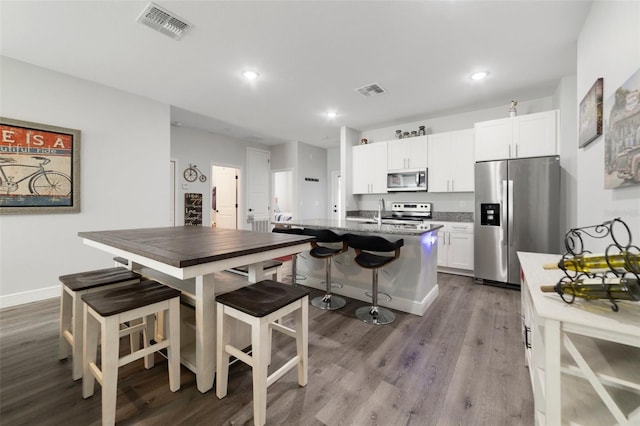 kitchen with a kitchen island with sink, stainless steel appliances, a kitchen breakfast bar, wood-type flooring, and white cabinets