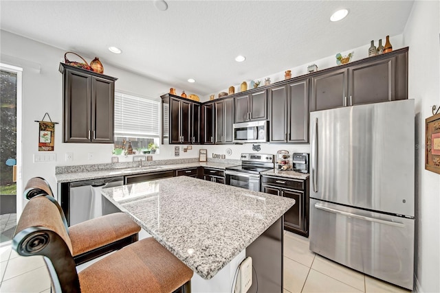 kitchen featuring dark brown cabinetry, light stone counters, a center island, light tile patterned floors, and appliances with stainless steel finishes