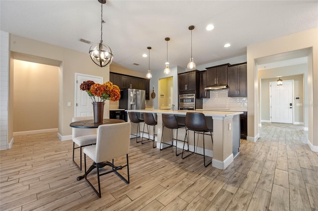 kitchen featuring a breakfast bar area, decorative backsplash, hanging light fixtures, a kitchen island with sink, and dark brown cabinets