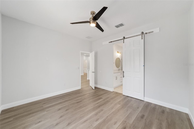 unfurnished bedroom with light hardwood / wood-style flooring, a barn door, ceiling fan, and ensuite bathroom