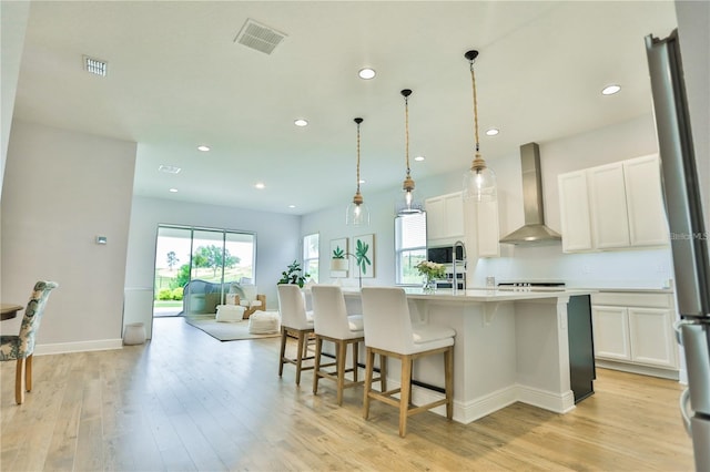 kitchen featuring wall chimney range hood, light hardwood / wood-style flooring, a kitchen island with sink, white cabinetry, and hanging light fixtures