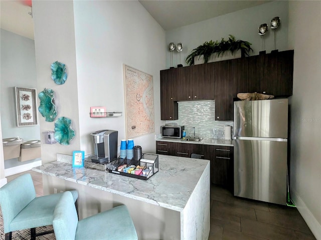 kitchen with stainless steel fridge, dark brown cabinets, light stone counters, decorative backsplash, and kitchen peninsula