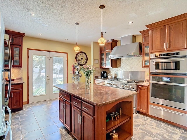 kitchen with french doors, wall chimney exhaust hood, hanging light fixtures, a kitchen island, and stainless steel appliances