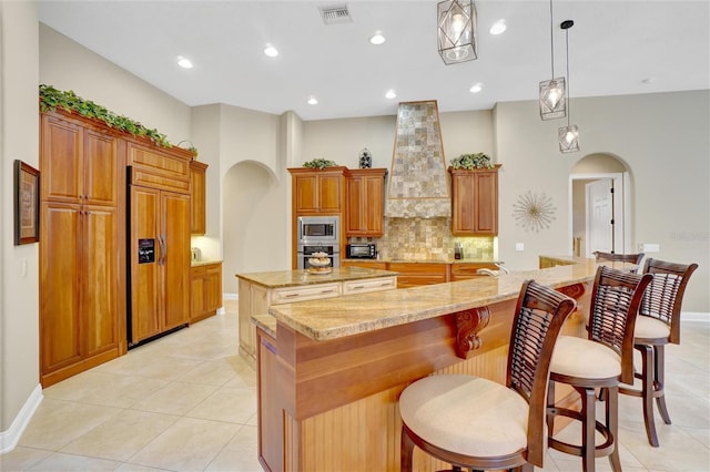 kitchen with a spacious island, premium range hood, light stone counters, built in appliances, and pendant lighting