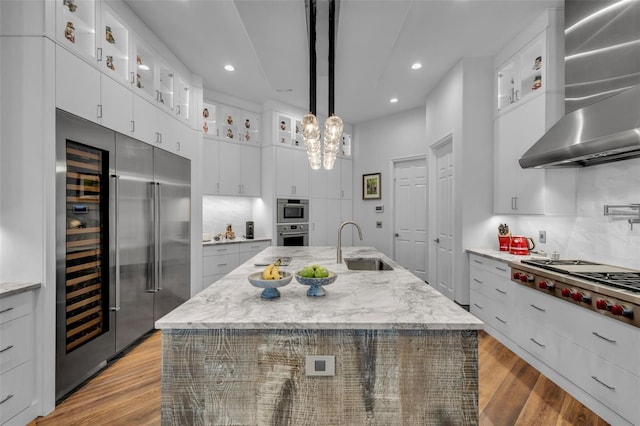 kitchen with wall chimney range hood, appliances with stainless steel finishes, an island with sink, white cabinets, and decorative light fixtures