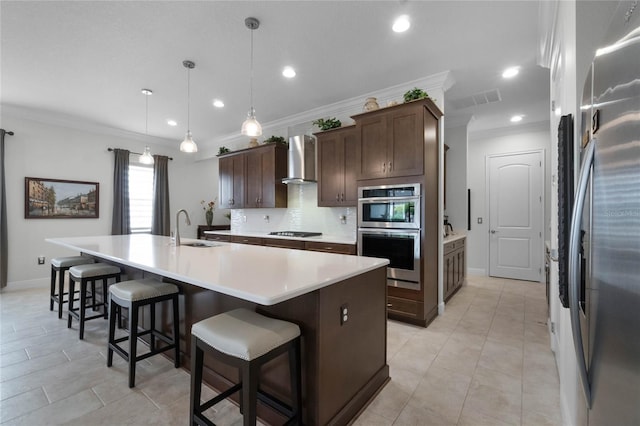 kitchen featuring wall chimney range hood, sink, appliances with stainless steel finishes, dark brown cabinetry, and a center island with sink